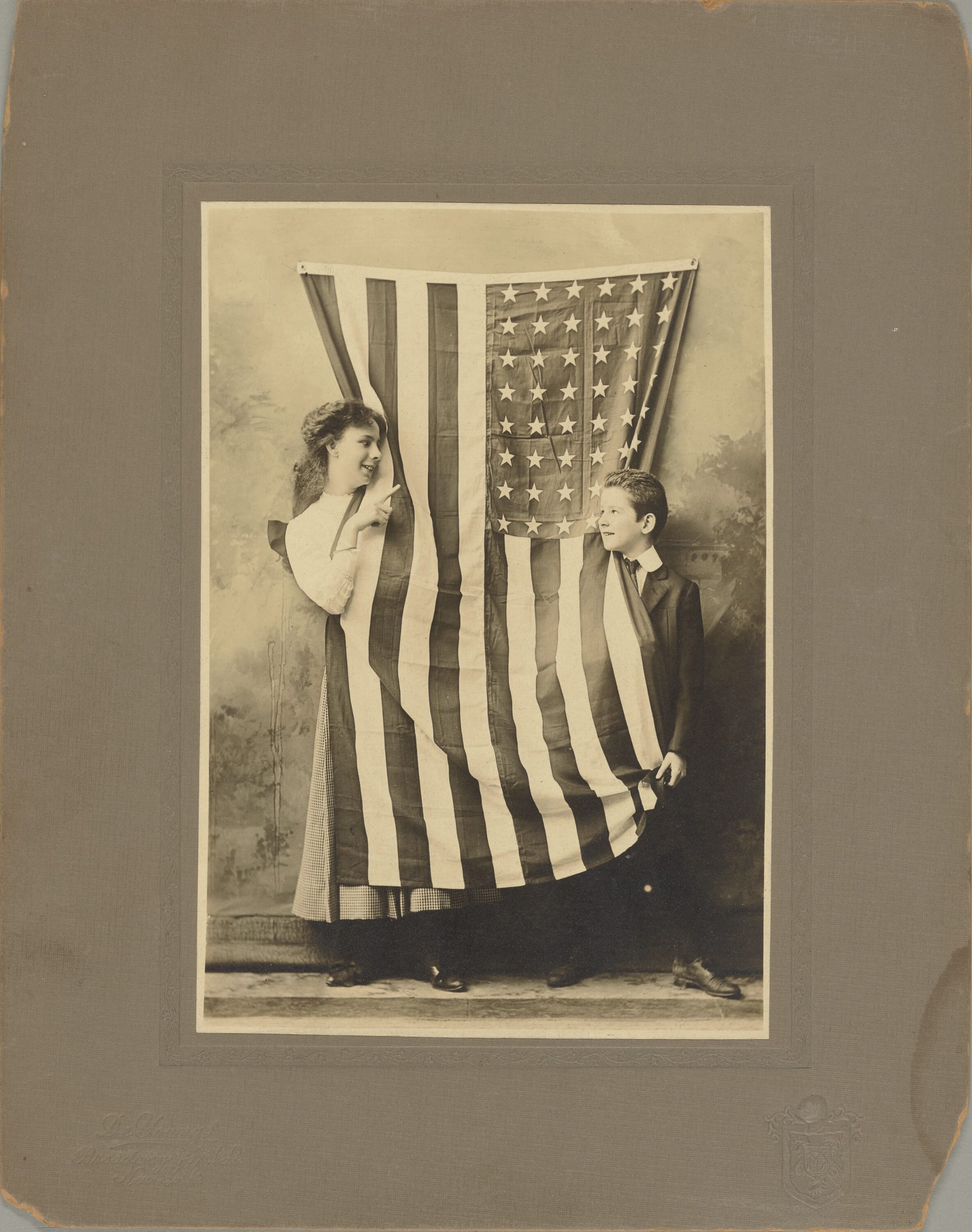 An old black & white photo of a girl and a boy draping themselves in an American flag.