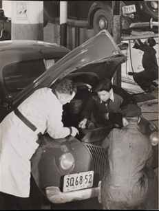 A group of mechanics look under the hood of an old car.