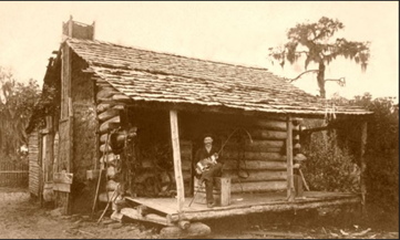 An image of a log cabin with a man sitting on the covered front porch.