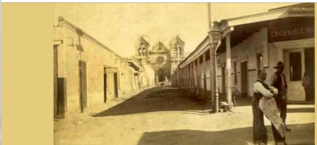 An old yellowed photograph of a street lined with single-storeyed buildings and a covered boardwalk. A church sits at the end of the street.