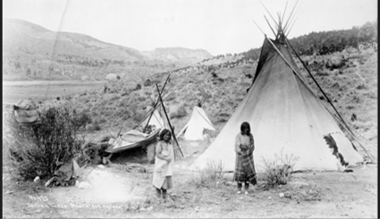 Two Apache women stand before a tipi camp amidst rolling hills. 