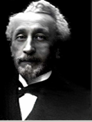 Photo of a gentleman with mustache, goatee, and receding grey hairline, dressed in formal attire with a continental tie.