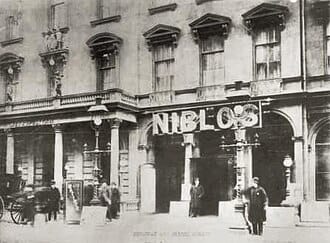 Horse carriages stand before a multi-story brick building with arched entrance, bearing the sign "NIBLOS".