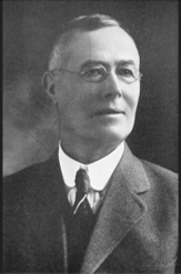 Photo of a gentleman in a suit and tie, wearing eyeglasses.