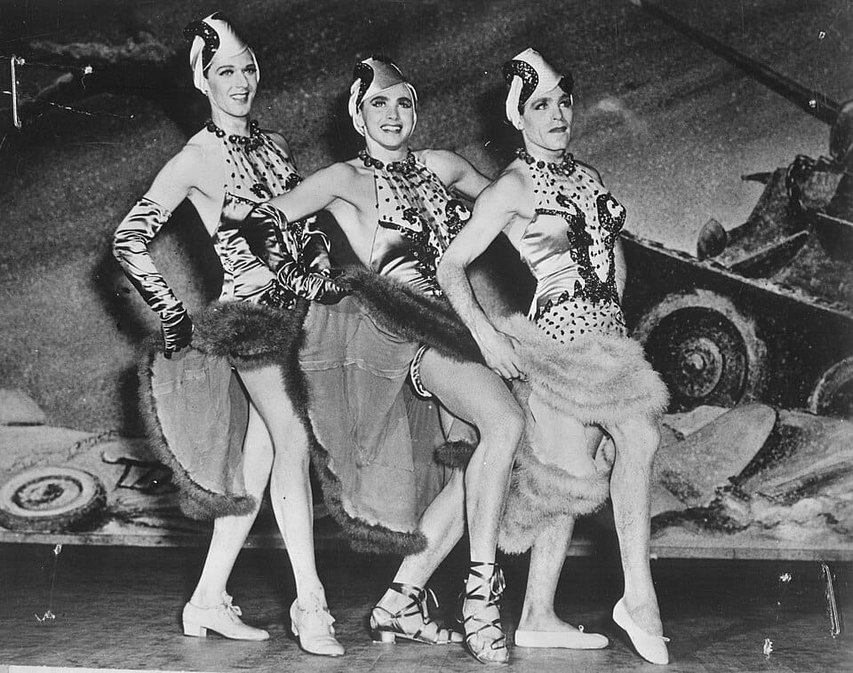Three men dressed as women dance in a broadway musical show, with a painted backdrop featuring an army tank.