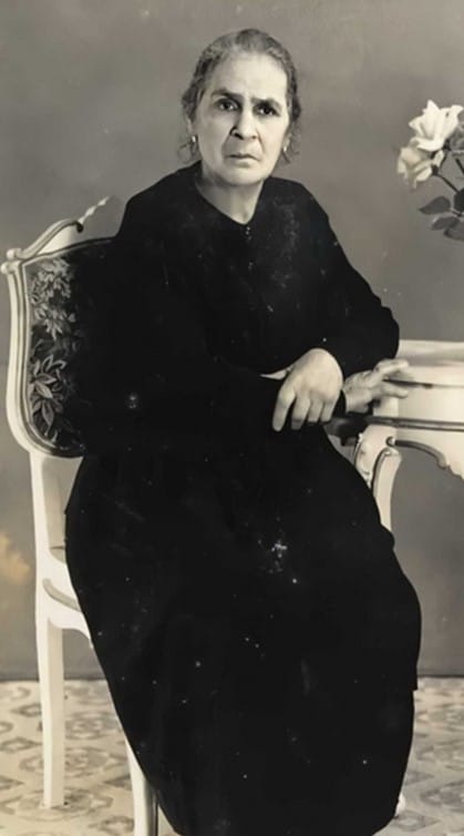 A portrait photo of an older woman in a long dark dress sitting at a table.