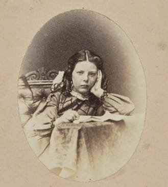 An unsmiling girl holds her hand to her face in this faded 1800s-era photograph with oval frame.