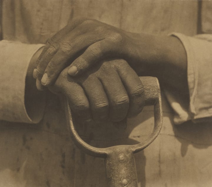 A photograph of a man's hands resting on the handle of a shovel.