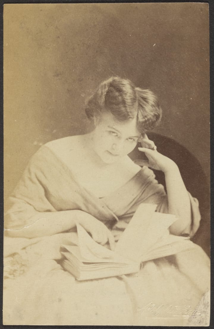 Portrait of a seated woman looking up from an opened book to which she's pointing.