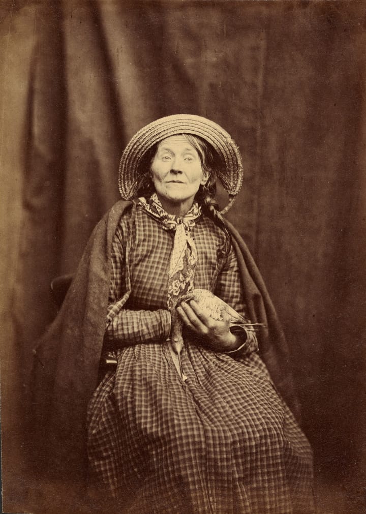 A sepia-toned photograph of a woman, in a long checkered dress and straw hat, seated holding a bird.