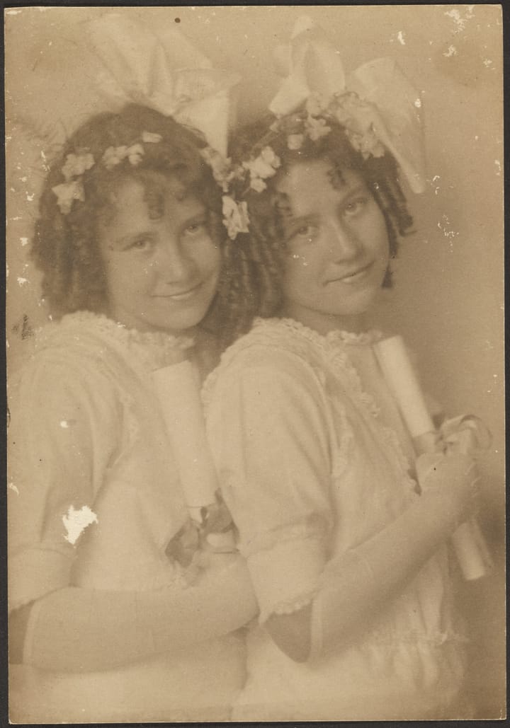 A old sepia-toned photograph of twin sisters, with ribbon and flower adorned curly hair, dressed in white.