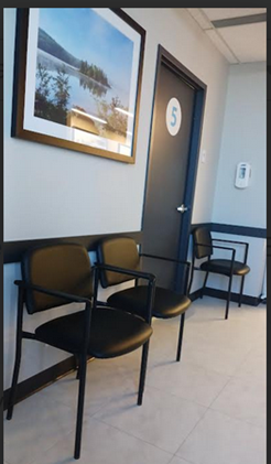 A waiting room with a couple of chairs against a wall, beneath a picture, with another chair on the other side of a doorway.